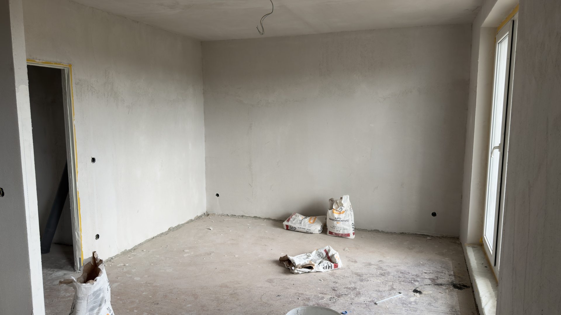 Wohnzimmer im Rohbau mit grauem Putz, blankem Estrich und offener Türöffnung